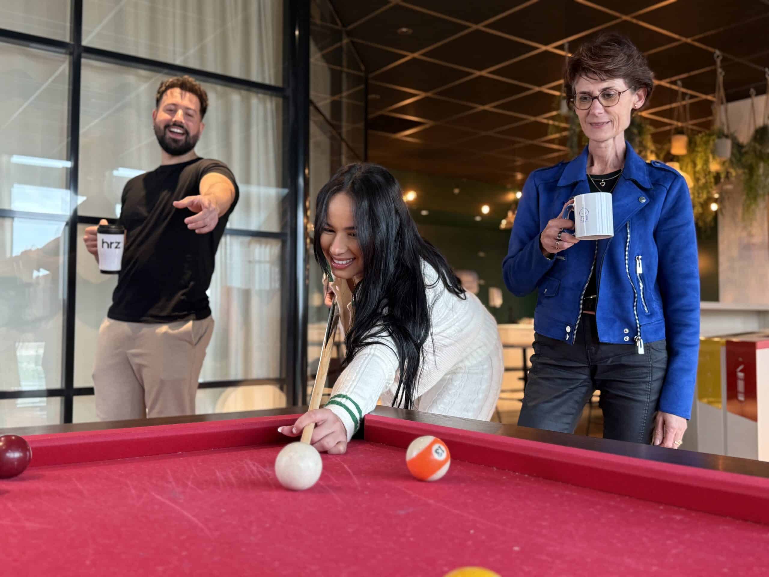 Trois collègues souriants jouent au billard dans un espace moderne. Une femme prend son coup. L'homme tient un mug marqué "hrz".