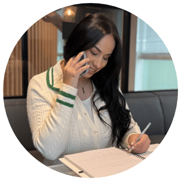 Communication efficace : prise de notes pendant un appel. Jeune femme souriante en gilet crème tricoté, écrivant dans un cahier pendant un appel téléphonique. Multitâche et prise de notes efficaces.