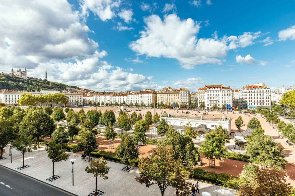 Place Bellecour Lyon : Parc et Basilique de Fourvière. Vue aérienne de la Place Bellecour à Lyon. Parc verdoyant, bâtiments classiques blancs, et basilique de Fourvière sur la colline sous un ciel bleu lumineux.