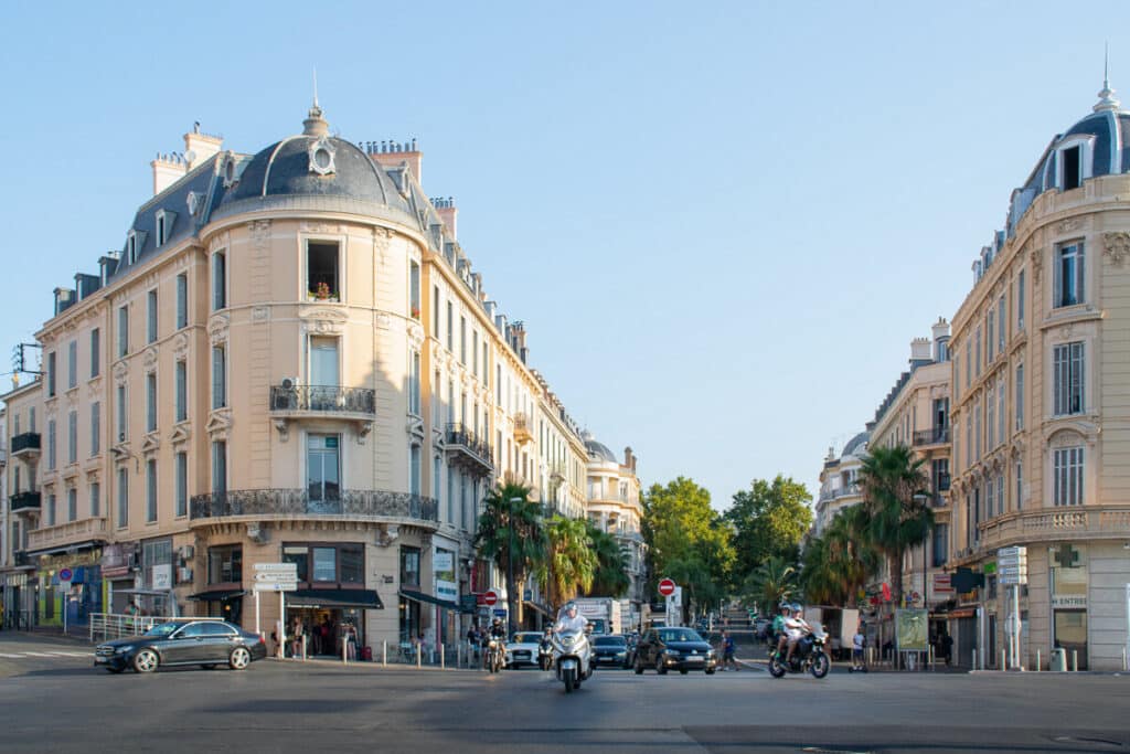 Architecture Haussmannienne : Carrefour ensoleillé d'une ville du sud. Intersection ensoleillée bordée d'immeubles haussmanniens beiges aux toits en dôme, avec palmiers et circulation de motos et voitures.