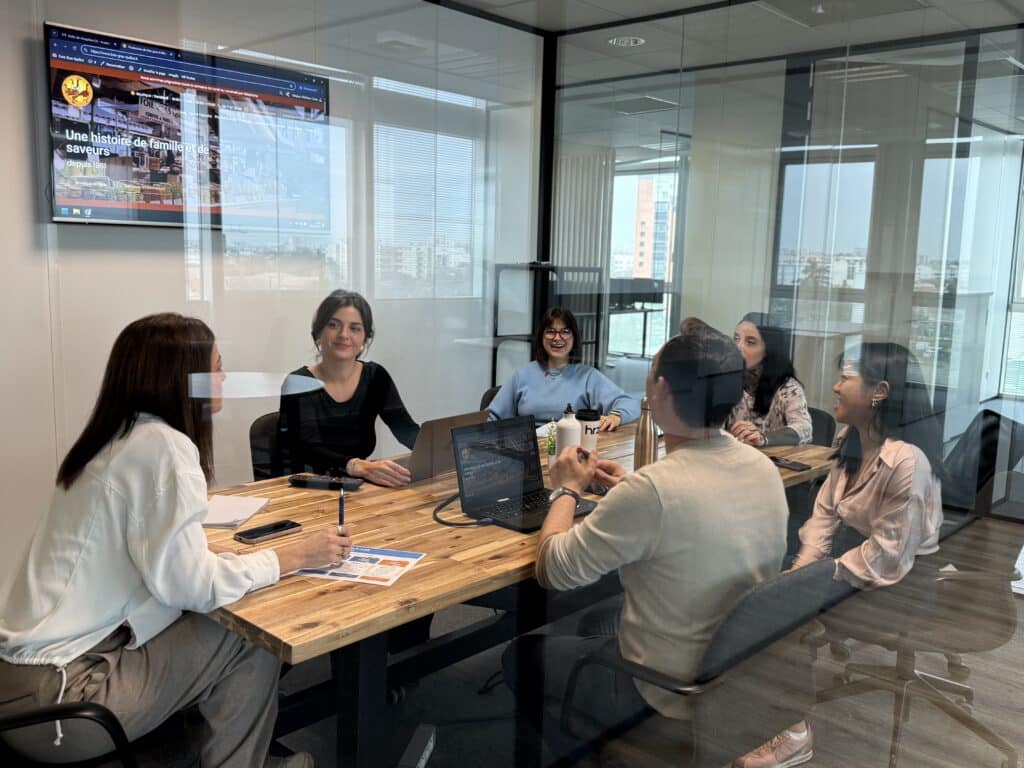 Equipe en réunion : Stratégie digitale et collaboration Réunion d'équipe souriante dans un bureau vitré. Un écran affiche le site "foie-gras-quillot.fr": Une histoire de famille et de saveurs.
