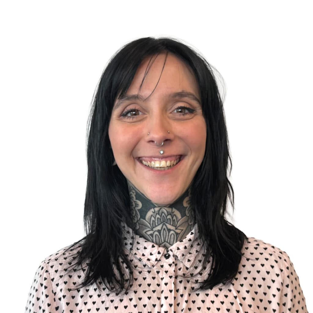 Portrait Femme avec Tatouage Cou et Piercings Visage Femme souriante aux cheveux noirs, tatouage mandala au cou et piercings faciaux en chemise à motifs cœurs.